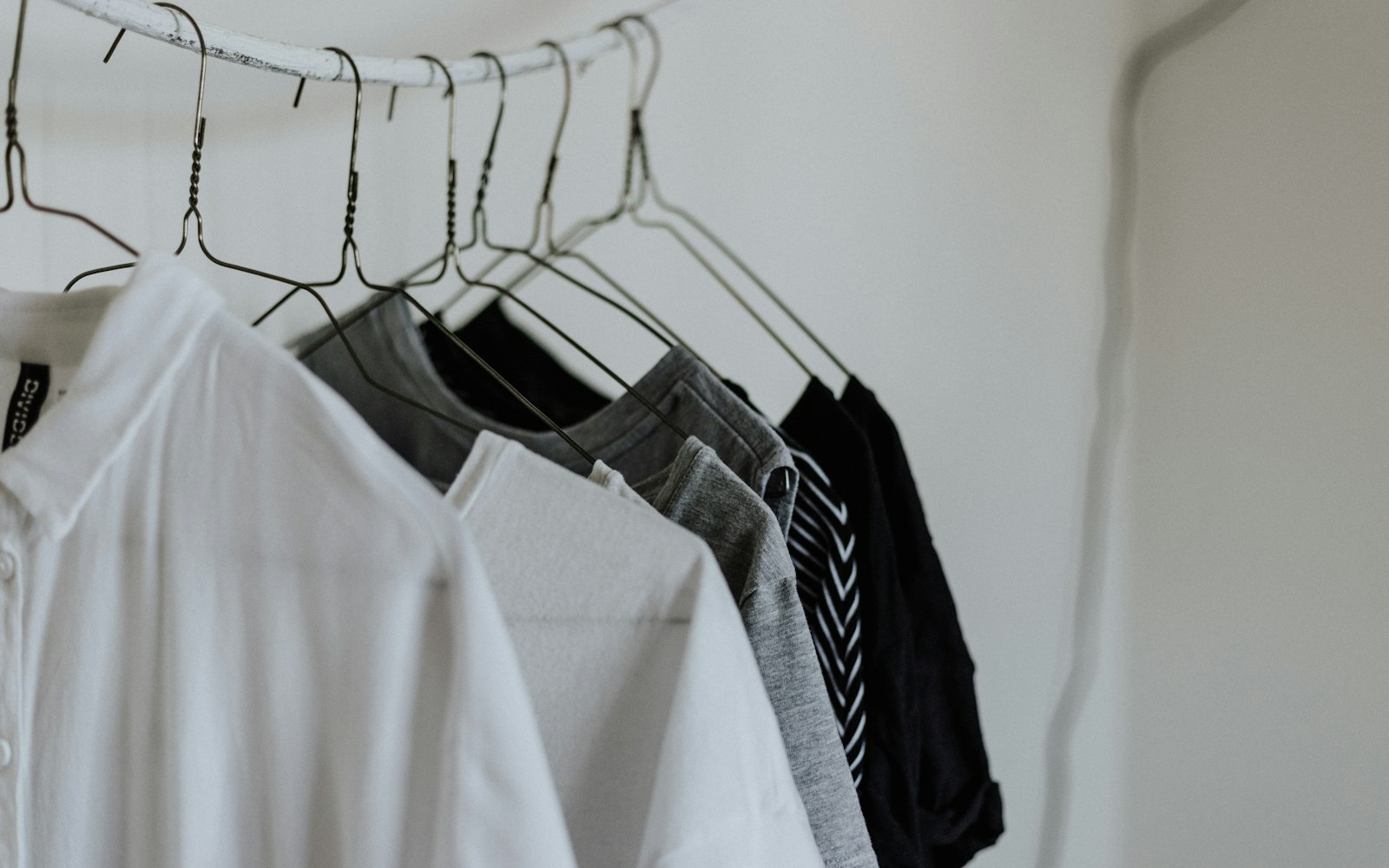 A row of plain white, grey and black garments on wire hangers on a rail against a white wall