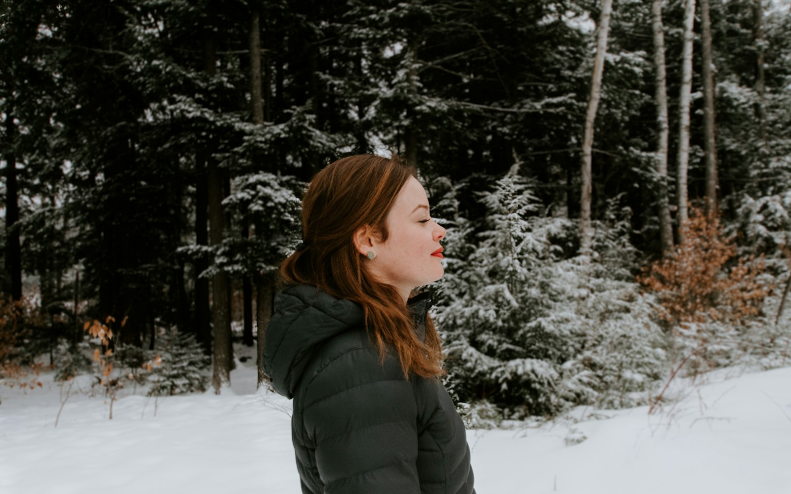A woman in a dark quilted puffer jacket standing in deep snow in front of a snow-covered pine forest