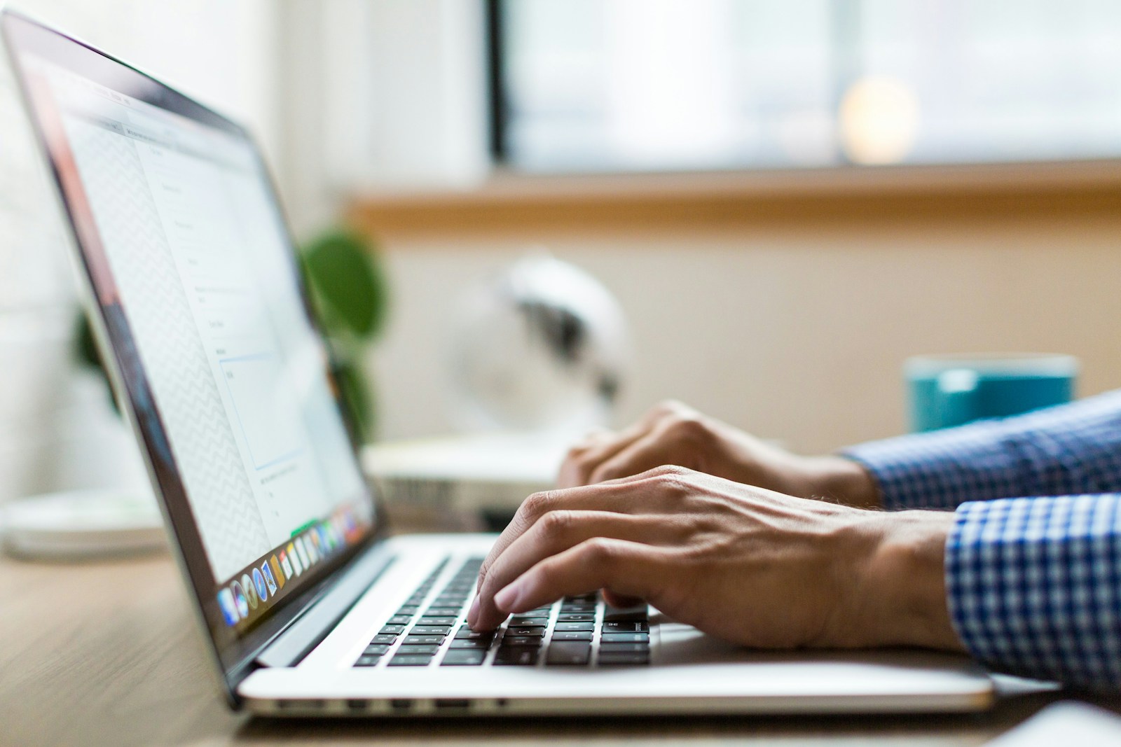 Person in a checked shirt typing on a silver MacBook at a desk